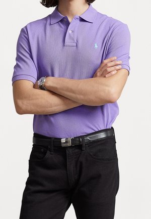 Person trägt ein violettes Poloshirt mit kleinem blauem Logo, schwarzen Gürtel, schwarze Hose, verschränkte Arme und eine silberne Armbanduhr am linken Handgelenk.