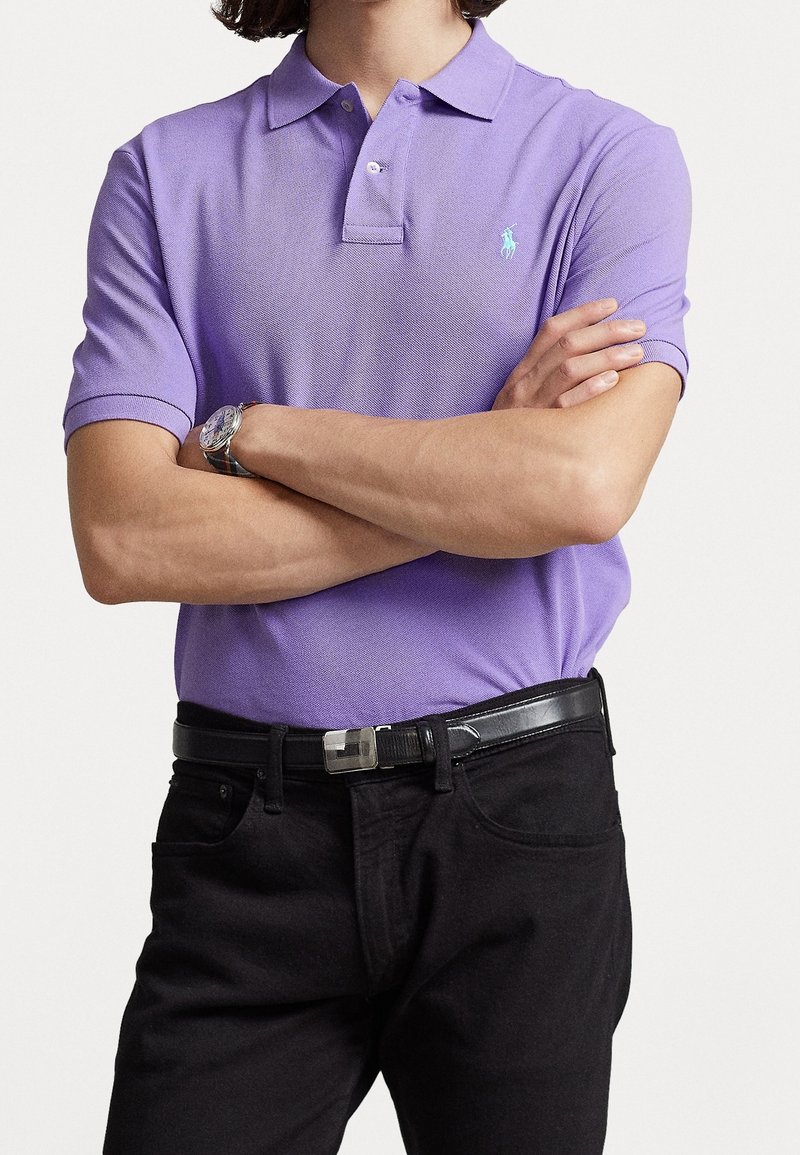 Personne portant un polo violet avec un petit logo bleu, ceinture noire, pantalon noir, bras croisés, et une montre-bracelet argentée au poignet gauche.