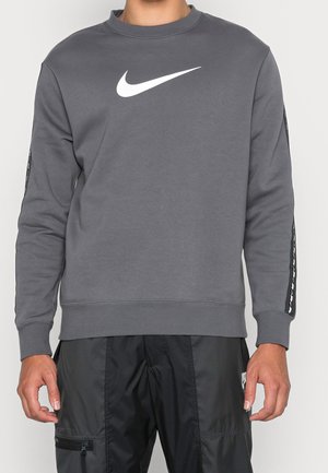 Mężczyzna w szarej bluzie Nike z dużym białym logo Nike swoosh na piersi i czarnych spodniach sportowych, stoi na jednolitym tle.