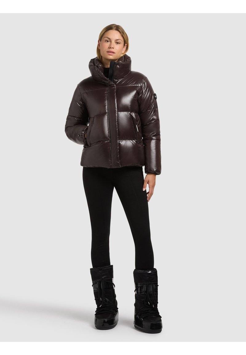 Puffig mörkbrun jacka med hög krage, glansig yta, knappar framtill och sidofickor, kombinerad med svarta leggings och snöskor.