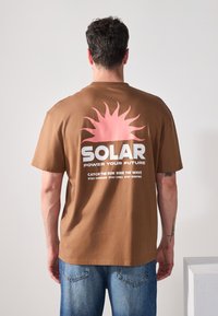 Brun kortærmet T-shirt med et stort rosa solgrafik og hvid tekst på bagsiden, med rund halsudskæring og løs pasform.
