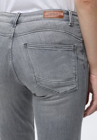 Gros plan d'une personne portant un jean en denim gris clair avec une poche arrière et un patch en cuir marron marqué ONLY à la taille.