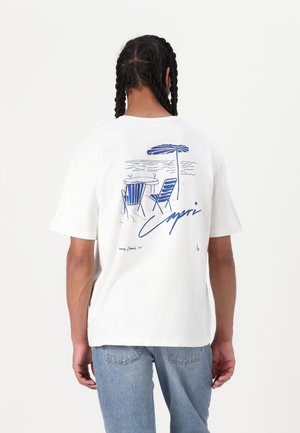White cotton t-shirt featuring a blue illustrated design of chairs and an umbrella, with "Capri" text and water background details.