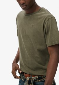 Camiseta de algodón verde oliva con cuello redondo, mangas cortas y un pequeño logo en el pecho, combinada con jeans azules y una camisa de cuadros atada a la cintura.