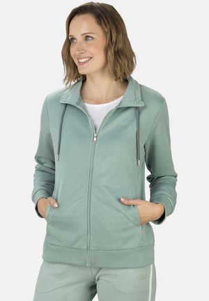 Frau lächelt, trägt hellgrünen Kapuzenpullover mit Reißverschluss und passende Jogginghose, Hände in den Taschen, schaut leicht nach links vor schlichtem Hintergrund.