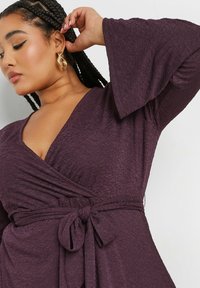 Vestido wrap de ciruela con una tela texturada, escote en V profundo y amplias mangas de campana, atado en la cintura con un cinturón a juego.