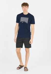 Mörkblå t-shirt i bomull med en vit våg- och fiskgrafik, kombinerad med svarta shorts och bruna slidesandaler.