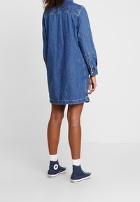 Denimklänning i blått, löst sittande med långa ärmar, knäppta manschetter och uppfälld krage, samexisterande med marinblå höga sneakers.