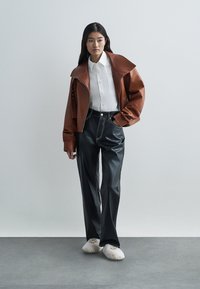 Veste en cuir marron avec col large, chemise blanche et pantalon en vinyle noir. Chaussures : chaussons en peluche blanc cassé. Design simple et moderne.