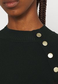 Haut en maille côtelée vert foncé avec un col montant et des boutons dorés décorant une épaule. Texture lisse et coupe ajustée.