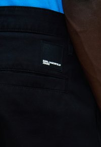 Πίσω όψη ατόμου που φοράει μαύρο τζιν Karl Lagerfeld και μπλε μπλούζα, με έμφαση στο λογότυπο του τζιν στην πίσω τσέπη.