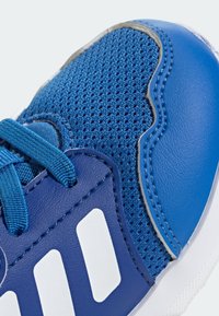 Zbliżenie niebieskiego buta sportowego, z siateczkowym wierzchem, syntetycznymi wstawkami, sznurówkami oraz białymi paskami po boku.