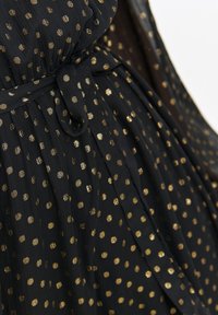 Czarna sukienka z luźnego materiału, zdobiona złotymi kropkami, z przewiązanym w pasie paskiem i plisowanymi detalami. Miękka tekstura z lekkim spadkiem.