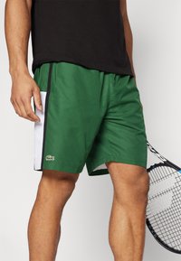 Shorts verts avec un panneau latéral blanc, ornés d'accents noirs et du logo Lacoste. Fabriqués en tissu léger, conçus pour le sport.
