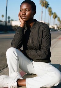 Jonge persoon met kort haar, gekleed in een zwart jasje en witte broek, zit op de stoep buiten, leunt met het hoofd op de hand en kijkt bedachtzaam.