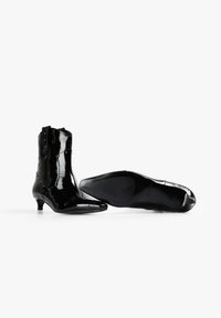 Botines negros de charol con punteras puntiagudas, tacones bajos tipo kitten y tachuelas decorativas en el lateral. Textura lisa y diseño elegante.