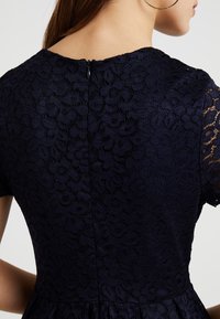 Námořnické krajkové šaty s krátkými rukávy, květinovým vzorem a diskrétním zipem na zadní straně. Texturovaná látka dodává hloubku.