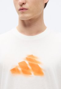 Maglietta bianca in cotone con un logo disegnato a spruzzo color arancione a forma triangolare sulla parte anteriore. Tessuto morbido e collo tondo classico.