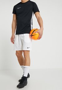 Persona con camiseta deportiva negra, pantalones cortos blancos y botas negras sosteniendo un balón de fútbol Nike naranja y negro frente a un fondo sencillo.