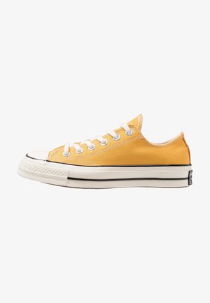 Żółty niski sneaker z płótna z białymi sznurowadłami, białą gumową podeszwą i czarnym paskiem, pokazany z profilu na białym tle.