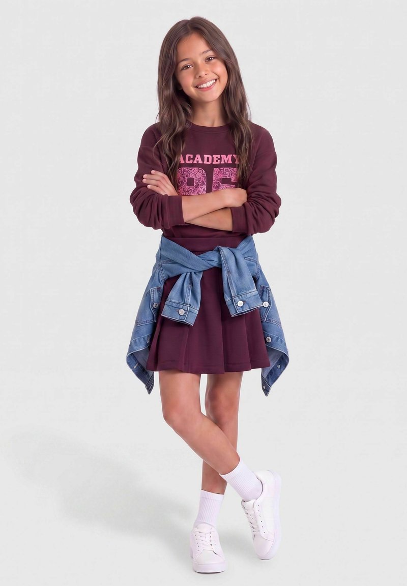Fille debout, bras croisés, portant une robe bordeaux, une veste en jean nouée autour de la taille, des chaussettes blanches et des baskets blanches, souriant à la caméra.