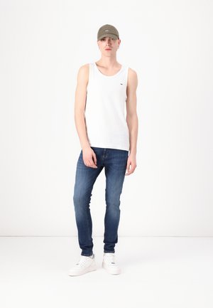 Joven con una camiseta sin mangas blanca, jeans azules, zapatillas blancas y una gorra verde oliva, de pie frente a un fondo blanco liso.