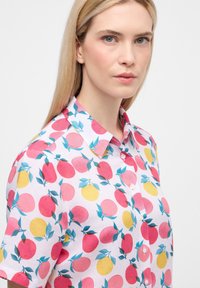 Camicetta a maniche corte realizzata in tessuto leggero; presenta uno sfondo rosa con motivi di frutta rossa e gialla e foglie verdi, colletto con bottoni.