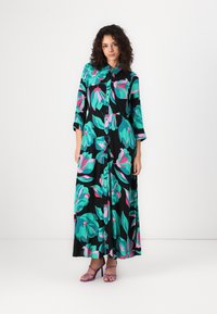 Robe maxi noire avec des motifs floraux vibrants verts et roses, à manches longues, avec une fermeture à boutons et une coupe ample. Associée à des talons strappy violets.