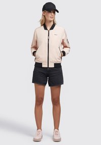 Lichtroze bomberjack met zwarte accenten, ritsluiting en zijzakken. Gemaakt met zwarte shorts en roze sneakers. Zwarte pet gedragen.