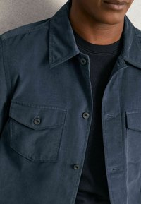 Marineblaues Cord-Jackett mit zwei vorderen Taschen und schwarzen Knöpfen, getragen über einem schwarzen Hemd. Der Stoff hat eine strukturierte Optik.