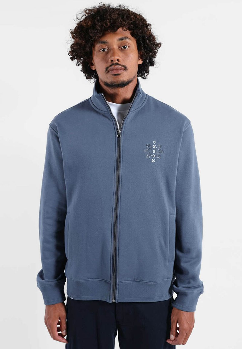 Homme aux cheveux bouclés portant une veste zippée bleue avec un petit logo brodé sur le côté gauche de la poitrine, debout devant un fond blanc.