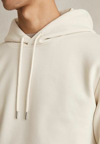 Krem hoodie napravljen od mekog materijala, s kapuljačom i vezicom s metalnim vrhovima te glatkim i teksturiranim završetkom.