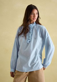 Femme aux longs cheveux foncés portant un chemisier bleu clair à volant sur le devant et un pantalon beige, debout devant un fond beige uni.