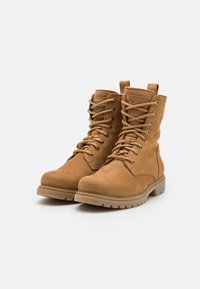 Botas em camurça bege com atacadores, gola acolchoada, ilhós de metal e sola de borracha resistente. Apresentam um logotipo na lateral e um cabedal texturizado.