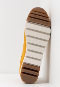 Semelle d'une chaussure jaune avec une épaisse semelle en caoutchouc blanc et marron rainurée, indiquant la taille 37, appuyée contre un coin blanc composé d’un mur et du sol.