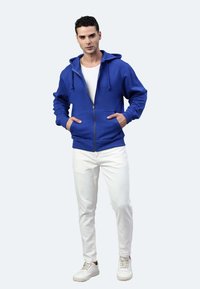 Felpa blu con zip, con due tasche anteriori, cappuccio regolabile con cordino e polsi ribattuti. Indossata sopra una t-shirt bianca e abbinata a pantaloni bianchi.