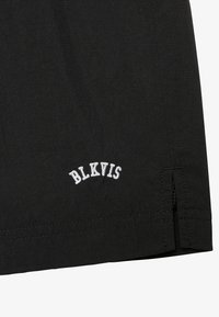 Pantaloni corti neri in tessuto presentano una superficie testurizzata, con il logo ricamato bianco "BLKVIS" sull'orlo. Il design include fessure laterali nella parte inferiore.