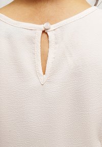 Svetle růžová blůza s texturovanou tkaninou. Má kulatý otvor na zádech zapínaný bílým knoflíkem. Jednoduchý design výstřihu.