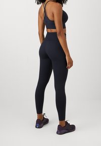 Ciemnoniebieski stanik sportowy z cienkimi, krzyżującymi się ramiączkami, w połączeniu z legginsami o wysokim stanie oraz prążkowanym wzorze, i ciemnymi sneakersami z purpurowymi akcentami.