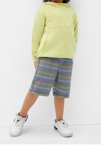 Světle zelená mikina s kapucí a přední kapsou, doplněná kostkovanými šortkami v modrých, zelených a hnědých odstínech. Bílé tenisky s černými Akcenty.