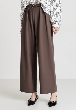Pantaloni larghi marroni con cintura dotata di una fibbia dorata a forma di D, abbinati a scarpe nere a punta. Tessuto liscio e fit sartoriale.