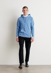 Sweatshirt azul claro com um bolso frontal, apresentando um cordão cinza e uma camisa branca por baixo. Combinado com calças e sapatos pretos.