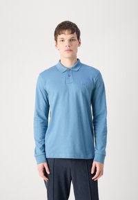 Langärmliges Polohemd in Hellblau, aus Baumwollmaterial, mit einem einfachen Kragen und einem kleinen Logo-Patch auf der Brust. Kombiniert mit dunklen Hosen.