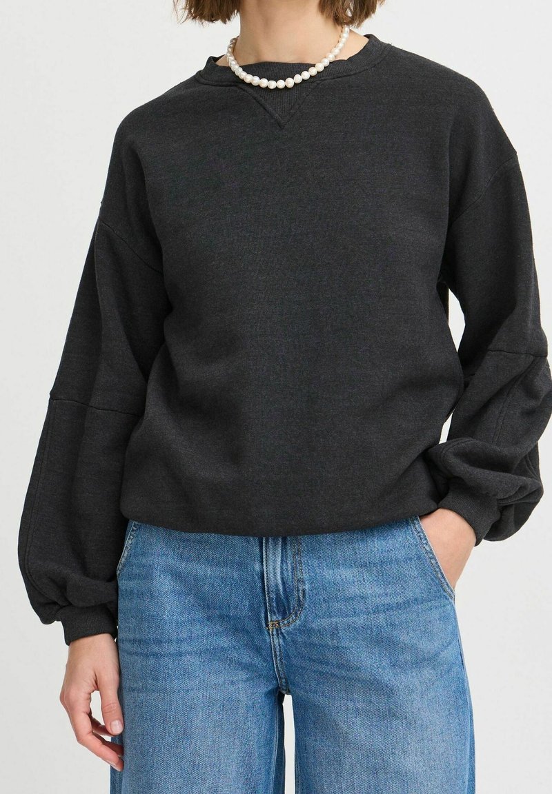 Personne portant un pull gris foncé à manches longues avec des manches bouffantes, un jean bleu clair et un collier de perles blanc, main dans la poche.