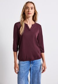 Top bordeaux à manches trois-quarts avec un col en V et un ourlet élastiqué. Tissu lisse avec une coupe décontractée. Associé à un jean en denim bleu.