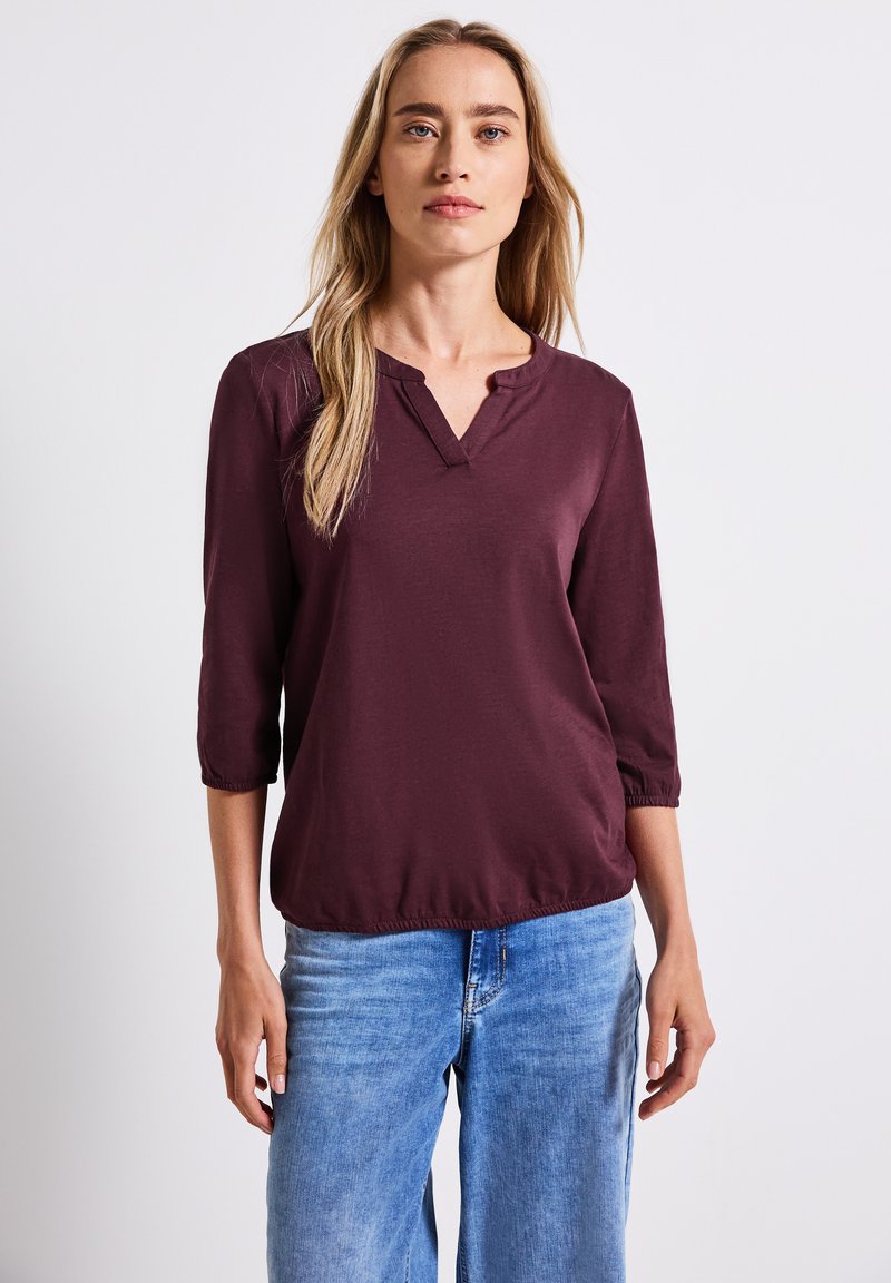 Top bordeaux à manches trois-quarts avec un col en V et un ourlet élastiqué. Tissu lisse avec une coupe décontractée. Associé à un jean en denim bleu.