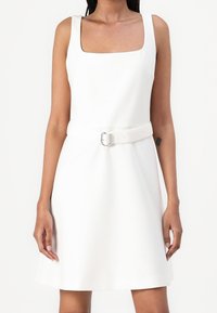 Vitt ärmlöst klänning med fyrkantig halsringning, åtsittande i midjan med ett vitt bälte och metallspänne, som har en utsvängd kjol.