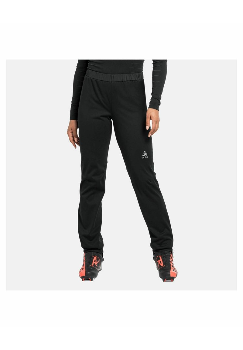 Personne portant un pantalon de sport noir et des chaussures de ski noires et rouges, debout devant un fond clair uni.