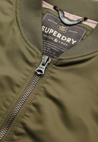 Oliwkowa bomberka z zamkiem błyskawicznym, charakteryzująca się gładkim nylonowym wykończeniem oraz subtelną etykietą w jodełkę od wewnętrznej strony.