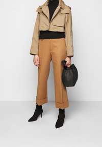 Giacca beige cropped, maglione nero a collo alto, pantaloni beige a gamba larga con risvolto, stivaletti neri a punta, mentre tiene una borsa nera texturizzata.